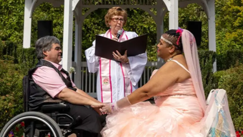 Marriage ceremony between two disabled people, one a white man in a wheelchair and the other a black woman in a fancy pink dress sitting down. An officiant stands between them.