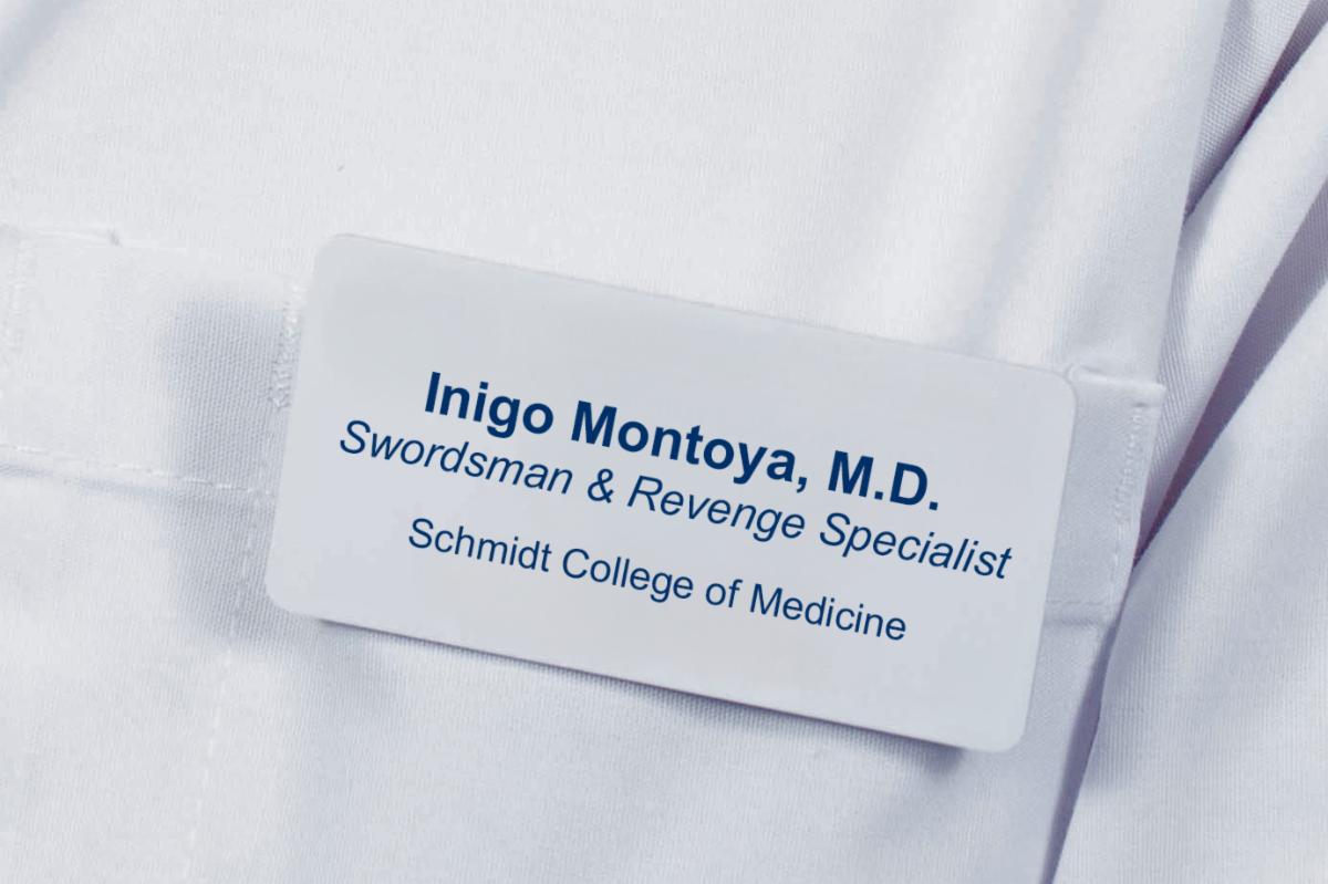 White name badge with Inigo Montoya, M.D. in bold blue text with his title of Swordsman & Revenge Specialist in italics. A new line with Schmidt College of Medicine