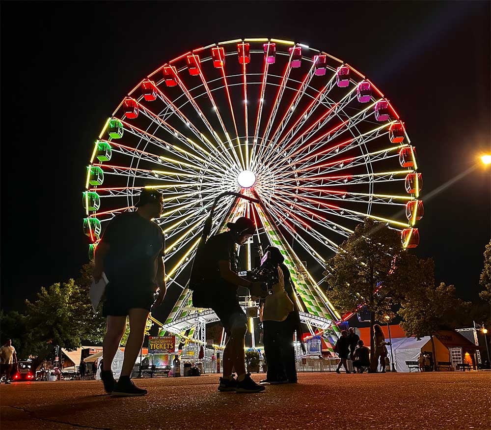 A film crew with cameras in front of the Great Big Wheel, a lit up ferris wheel, at night.