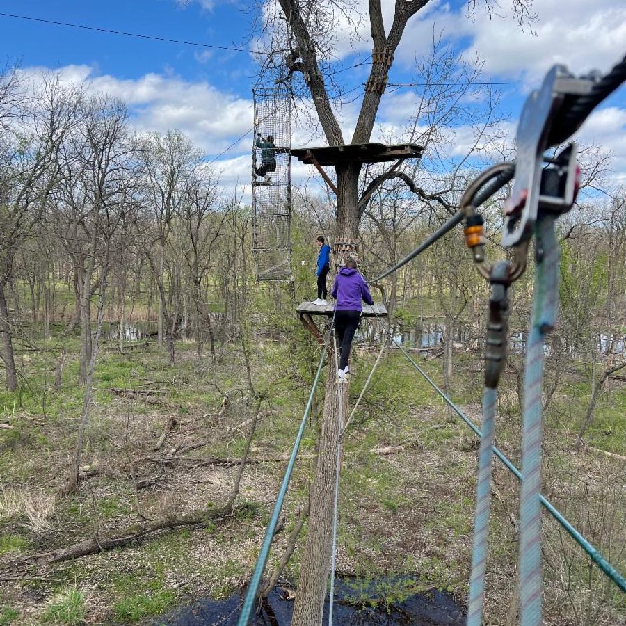 A student balances on a ropes course with treetops in the background