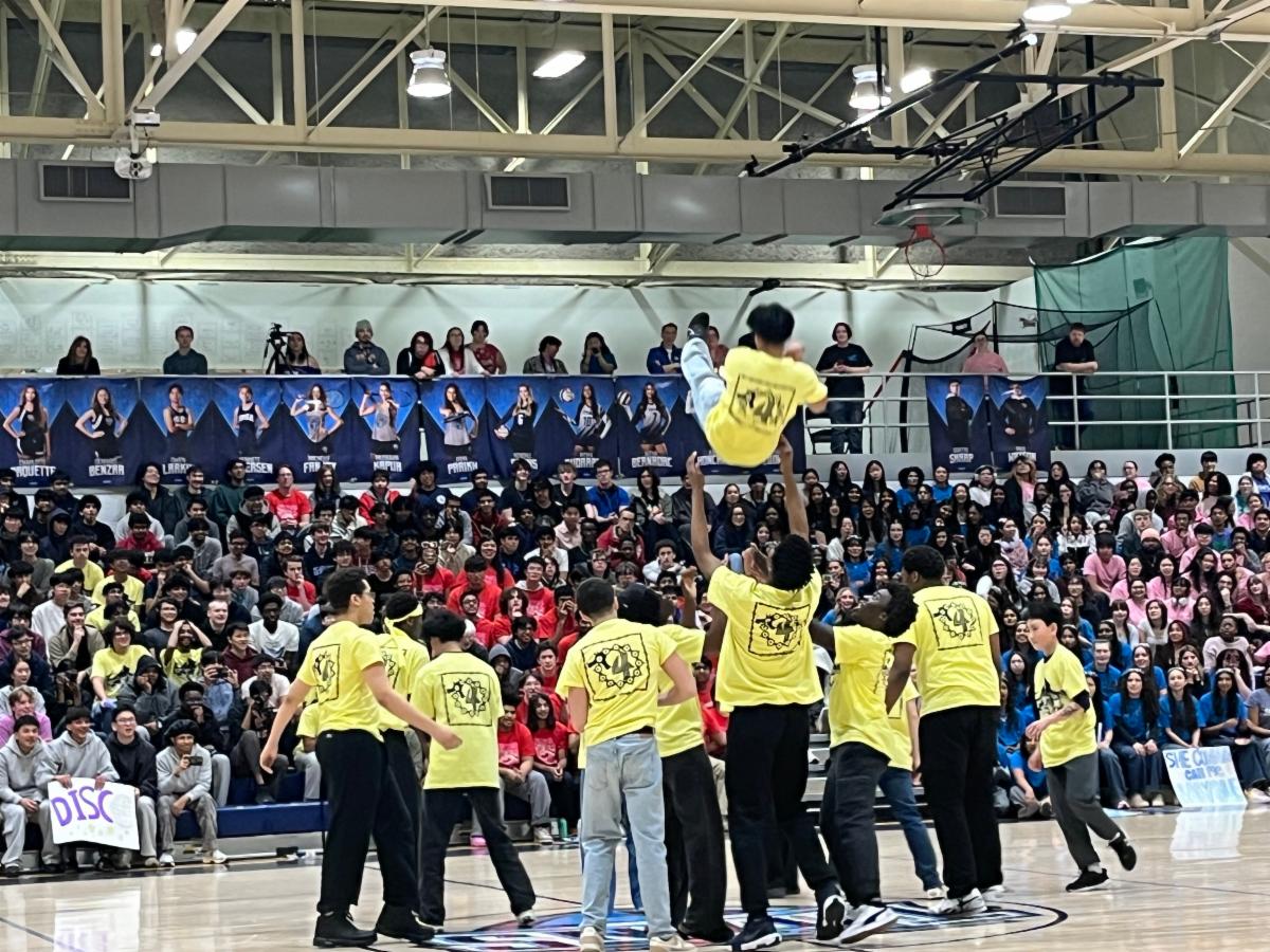 A group of students in yellow t shirts throw a classmate into the air in a cheerleading stunt while students on the bleachers cheer them on