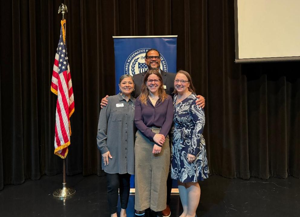 Four adults pose smiling in front of a banner featuring IMSA's crest on a stage. 
