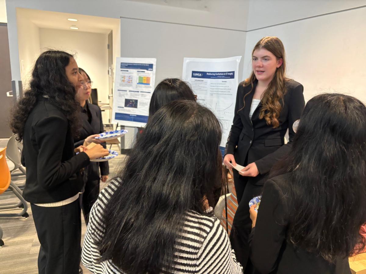 A student in a suit stands in front of her research poster presented to a group of girls. 