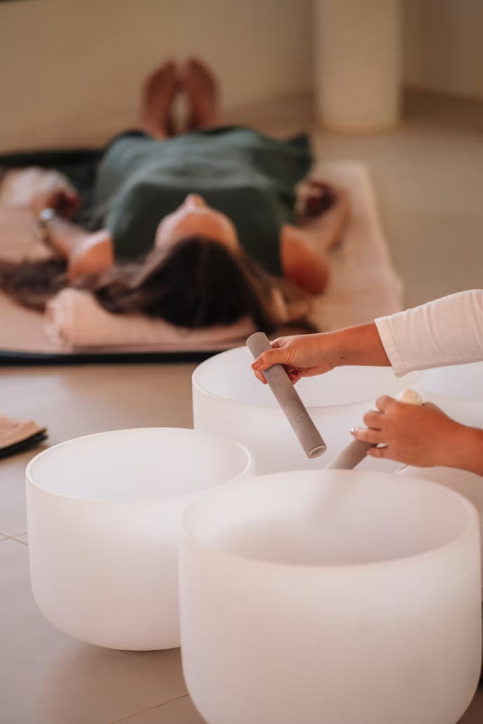Close up image of hands holding a mallet hitting a sound bath bowl while someone lays in meditiation in the background