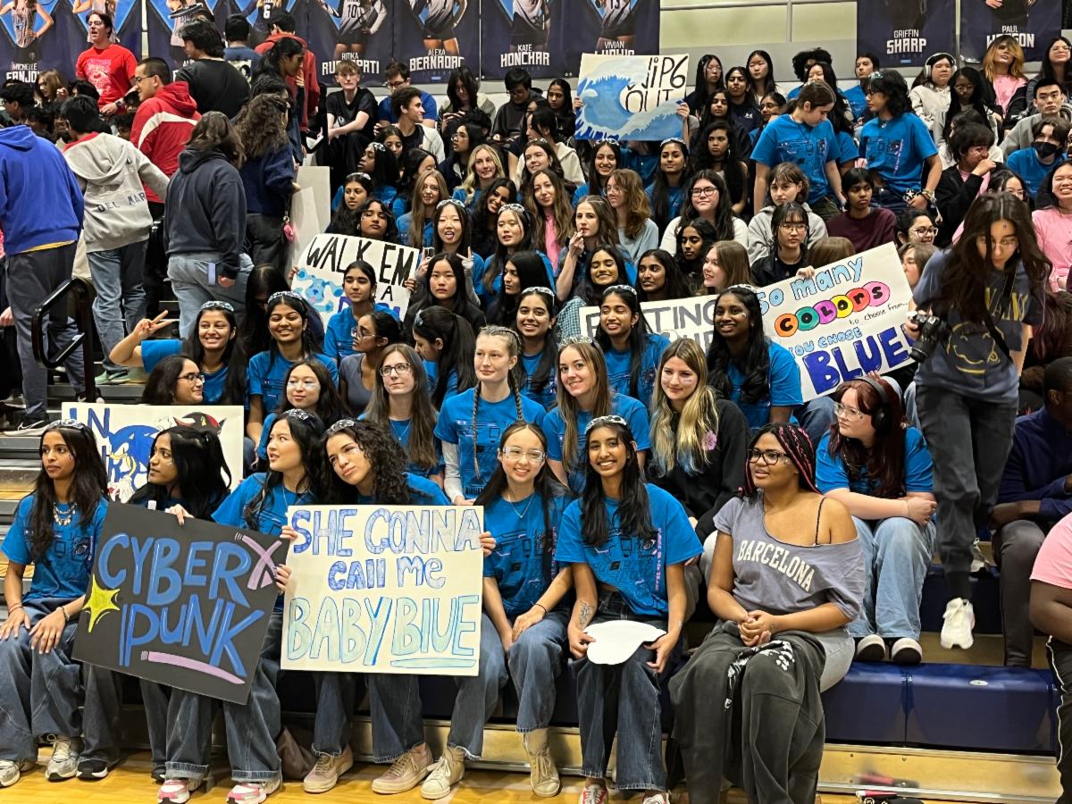 A group of students in blue shirts sit on bleachers smiling and holding up signs supporting their classmates
