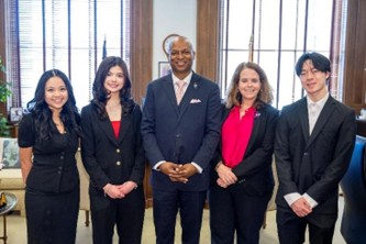 Five people stand side by side in an office setting, including three students in formal black attire and two adults in professional clothing. They are smiling and posing in front of large windows and wooden furnishings.