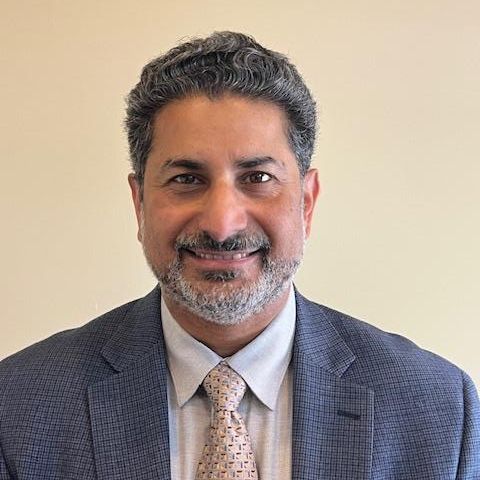 A man with dark hair and a goatee smiles in front of an off-white background. He is wearing a grey suit jacket and cream colored tie.