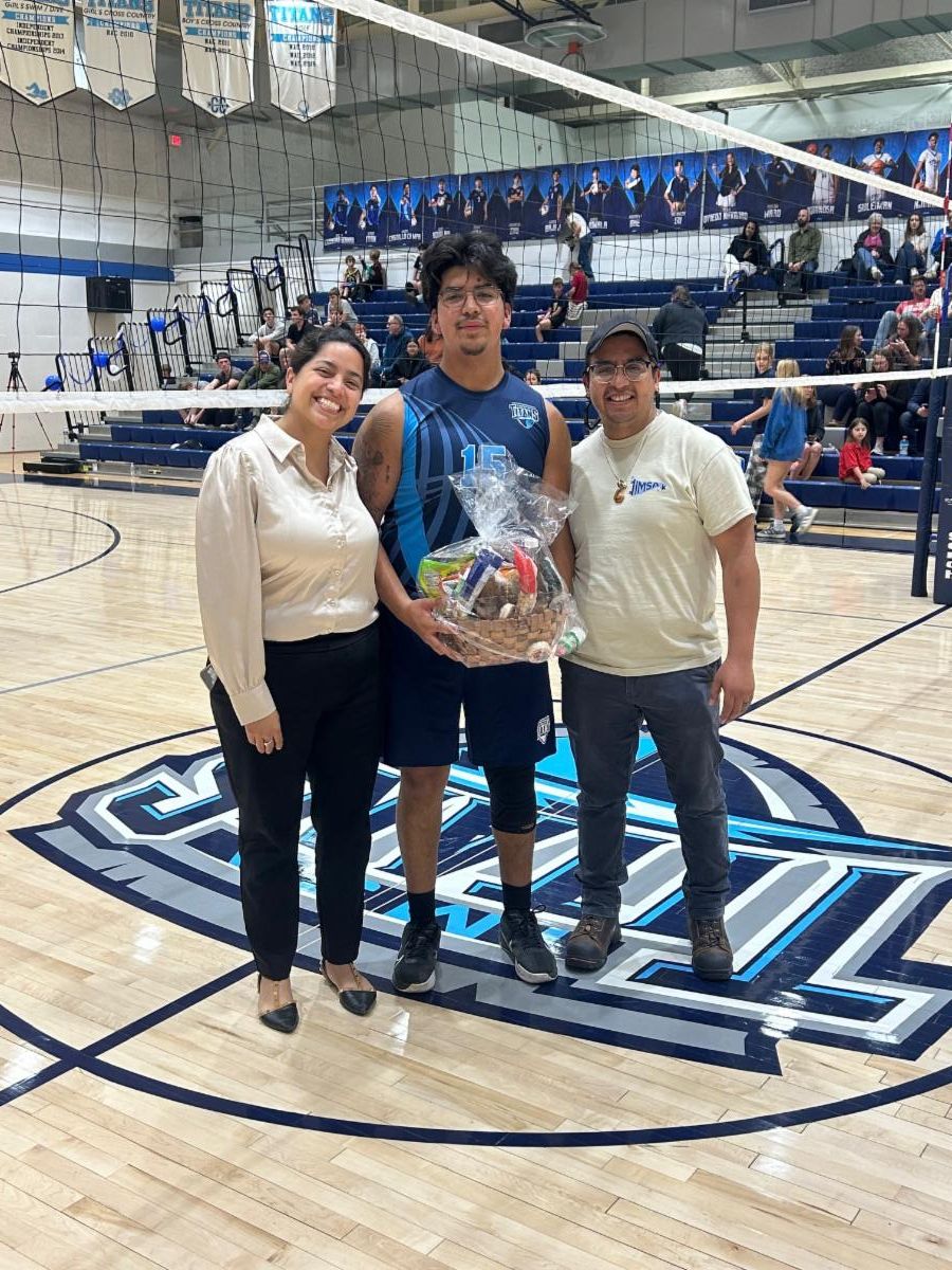 A senior boys volleyball player poses with his parents on senior night holding a gift basket near a volleyball net