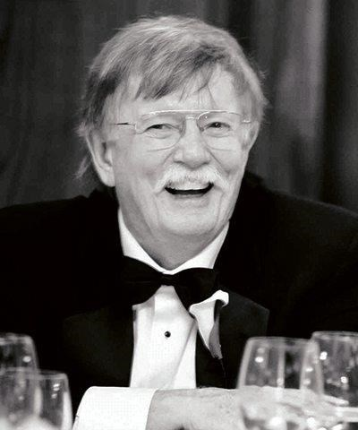 A black and white picture of a man with grey hair, a mustache and glasses laughs at a banquet table while wearing a tuxedo 
