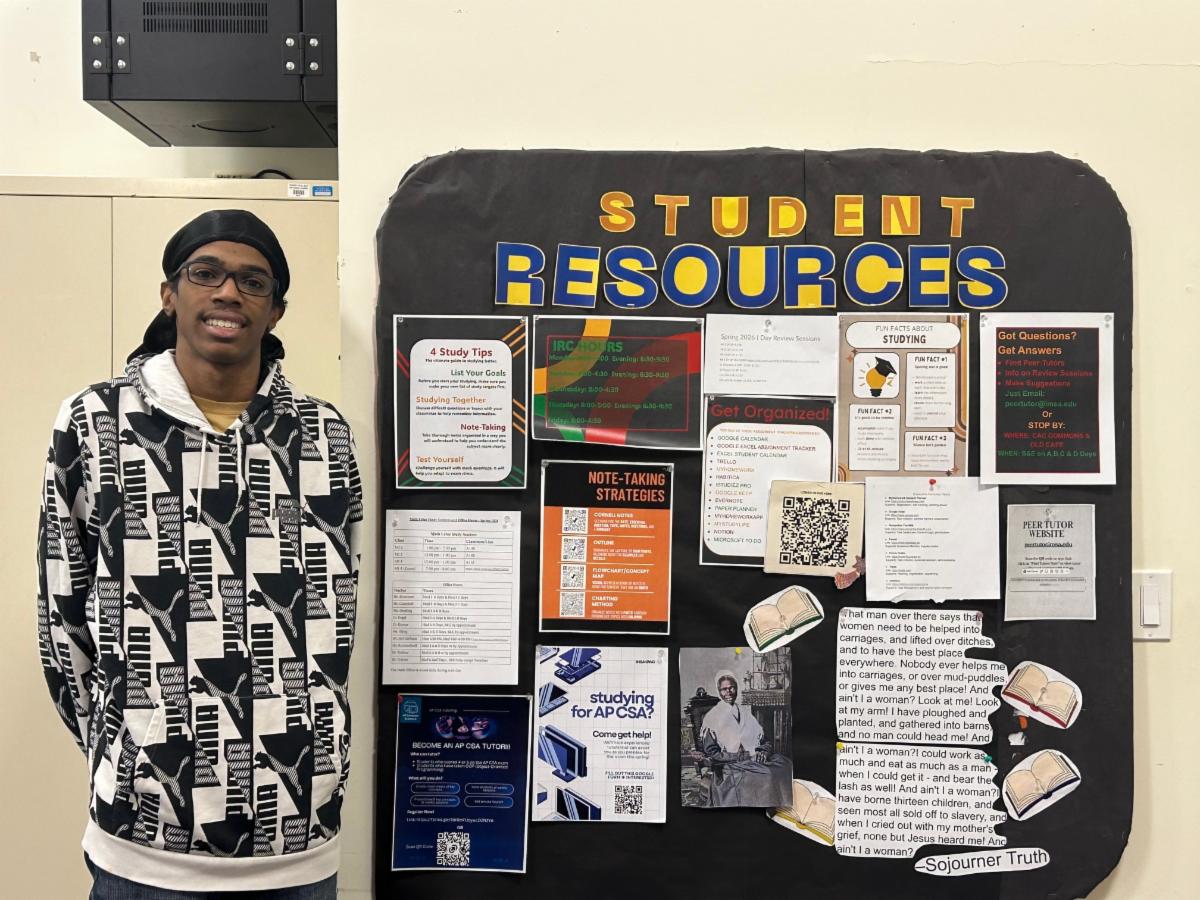 A student a black-and-white patterned hoodie stands smiling beside a bulletin board titled âStudent Resources.â The board displays colorful flyers with study tips, note-taking strategies, tutoring information, sc