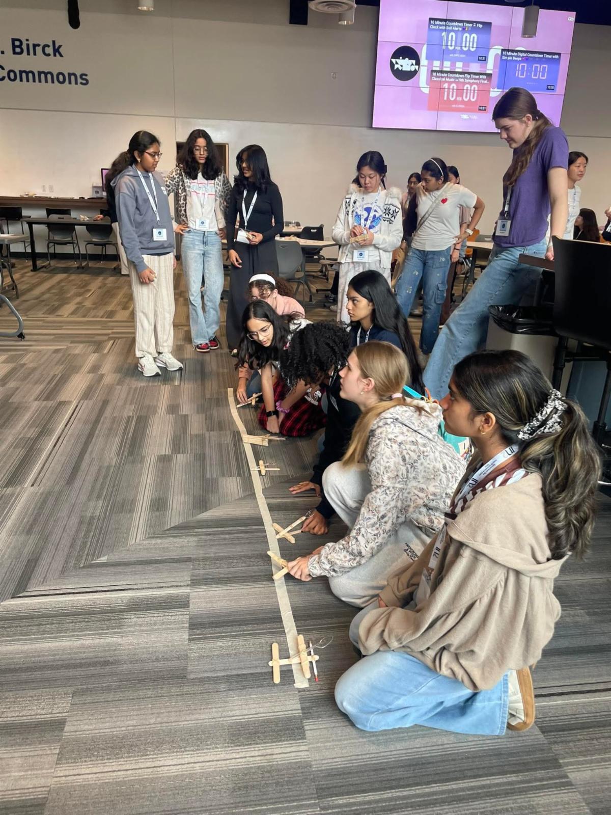 A group of girls kneels on the floor lining up small homemade catapults while other girls watch