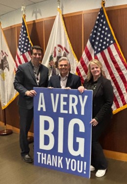 Three people stand together holding a large blue sign that reads "A Very BIG Thank You!" American and Illinois state flags are displayed on flagpoles in the background.
