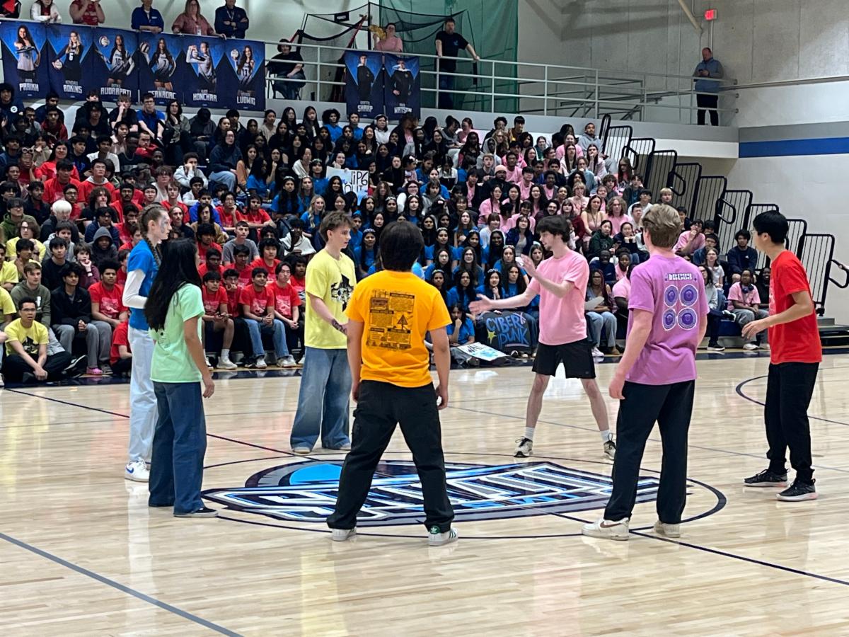 Students in multi colored shirts stand in a circle while a crowd of students on the bleachers cheer them one