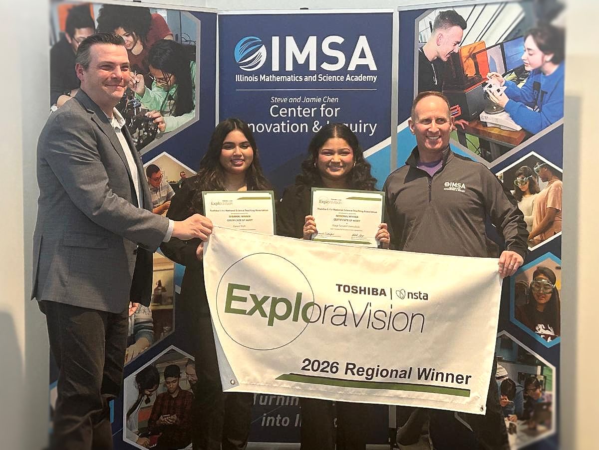 Two girls stand behind a banner that reads "exploravision" as they hold up two award certificates. Two men in business casual attire hold up the banner for them and smile for the photo.