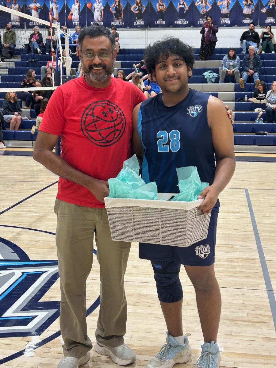 A senior boys volleyball player poses with his parents on senior night holding a gift basket near a volleyball net