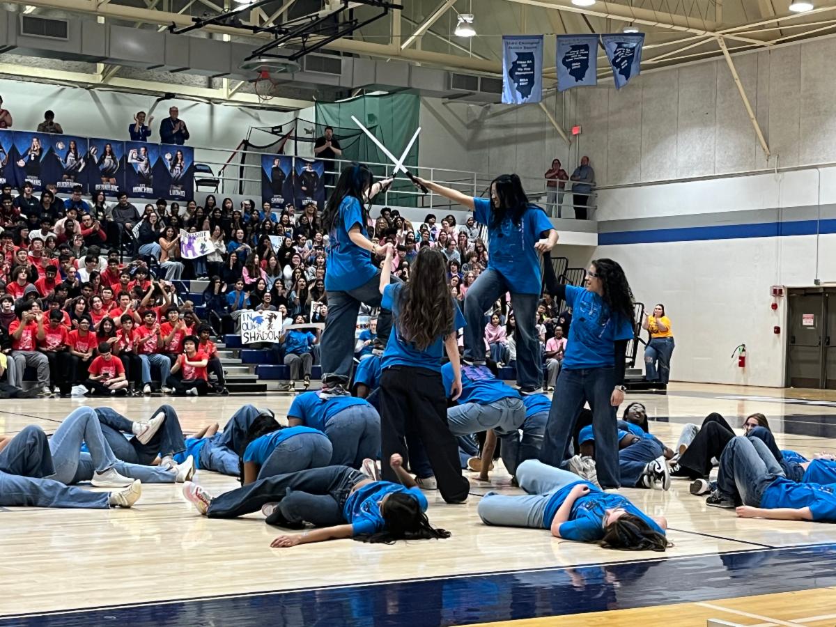 A group of students in blue t shirts form a human pyramid in a gym while students on the bleachers cheer them on