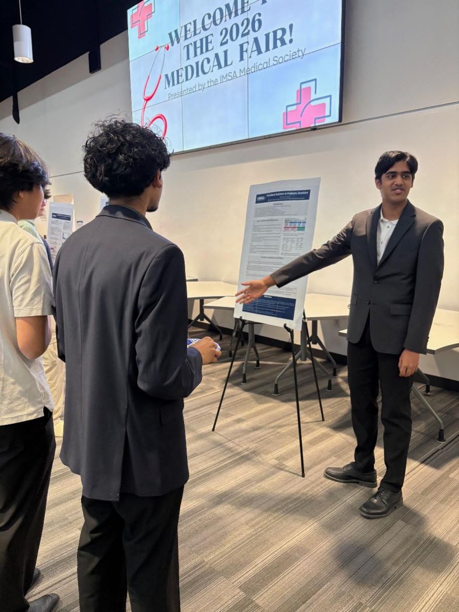 A student presents a research poster to peers at the 2026 Medical Fair beneath a welcome screen.
