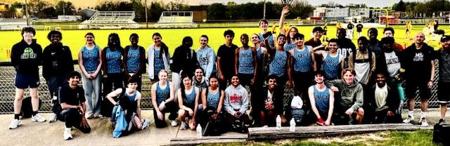 A group of students in track and field uniforms pose for a picture at dusk outside at a track meet.