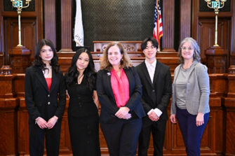 Five individuals pose inside a courtroom, standing in front of a judgeâs bench with American flags visible. Three students in formal attire stand alongside two adults, all dressed professionally and smiling.