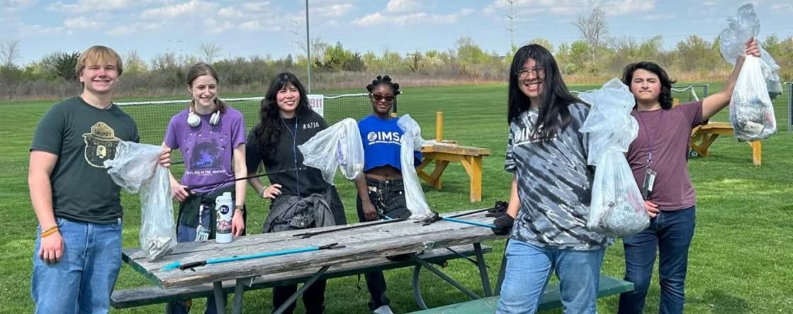 Students stand around a picnic table outside on a sunny day with blue skies and clouds