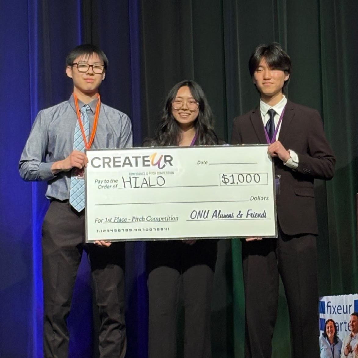 Three students dressed in business casual attire hold up a giant check for winning first place at the pitch competition.