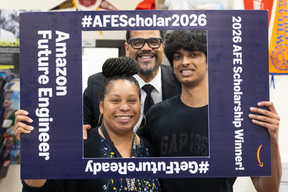 A student and two adults smile and pose in a carboard photo frame with the words Amazon Future Engineer on it. 