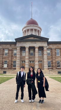 Three students stand on a walkway in front of a large historic building with a domed roof. All three are dressed in professional attireâtwo in dark suits and one in a black outfit with a red topâposing together outdoors under a cloudy sky.