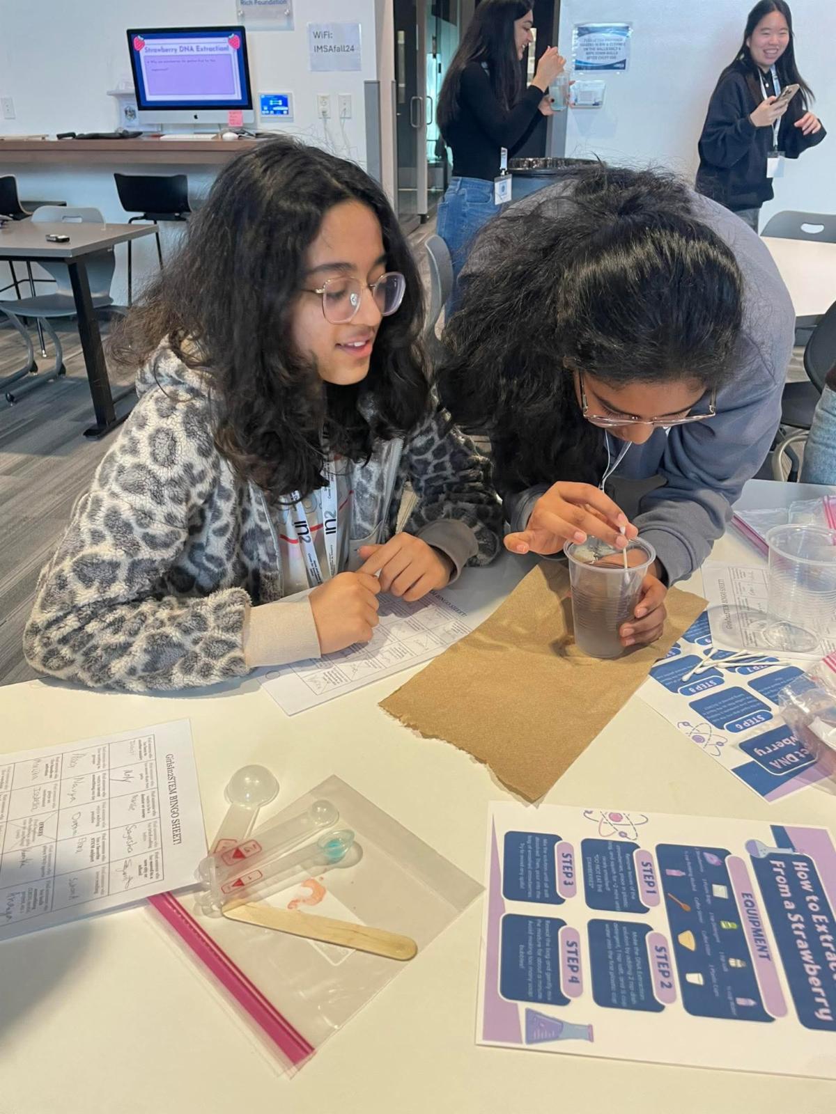 Two girls lean over the table observing a DNA extraction activity in a cup of liquid