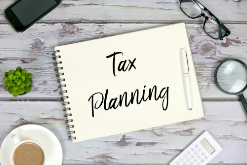 Top view of mobile phone_plant_coffee_calculator_magnifying glass_sunglasses_pen and notebook written with Tax Planning on wooden background.