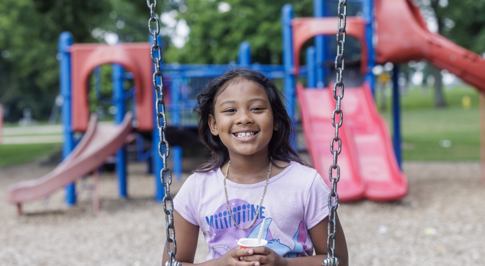 Aneah sits on a swing in the park, snacking on a cup of yogurt.