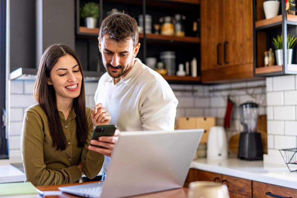 Young smiling couple using laptop and smartphone checking their home finances in the kitchen