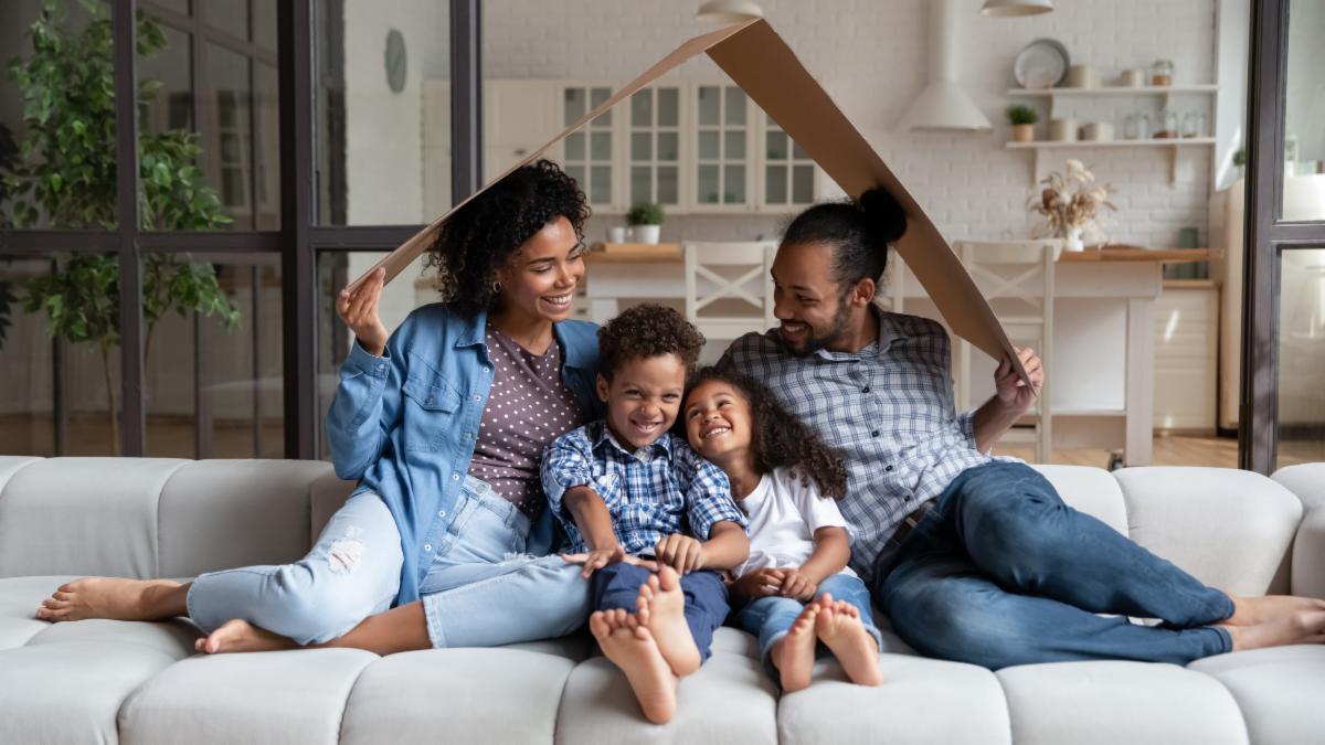 Happy affectionate young African American couple parents sitting under carton roof on comfortable couch with small adorable children son daughter celebrating moving into own home real estate concept