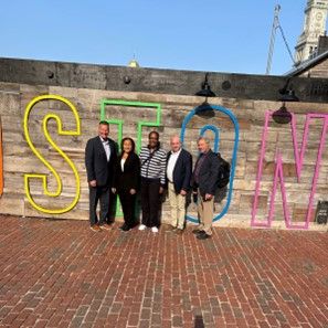Chair Bryan Albrecht, Eva Mitchell, Vice-Chair Annette Parker, Treasurer Boyd Worsham, and Scott Solberg stand in front of a "Boston" sign.