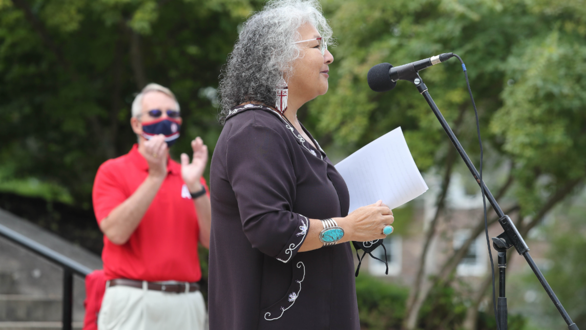 Elder Lorraine Whitman address students from the podium. President Peter Ricketts looks on and applauds.