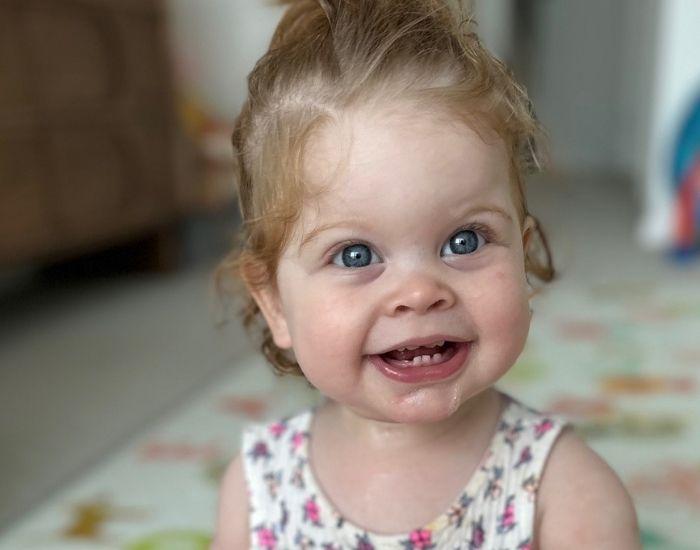 A smiling toddler with curly, light hair wears a floral dress. The background is softly blurred, conveying warmth and cheerfulness indoors.