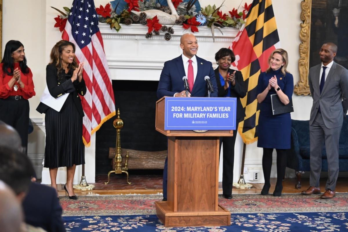 Maryland Governor Moore stands behind a podium with a sign that reads, " 2024:  Maryland's Year for Military Families." Five people in business attire applaud behind him.