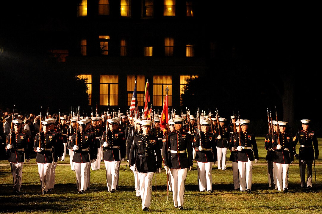 An evening parade of United States Marines in formal uniform across a greenspace.