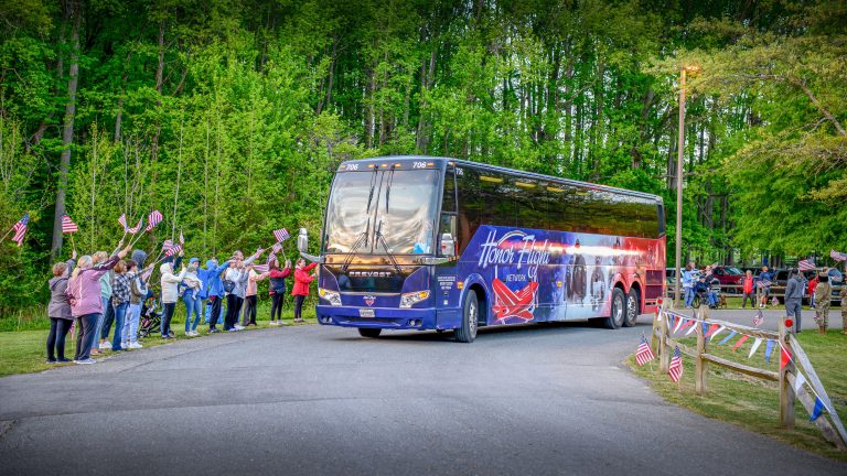 A brightly colored tour bus makes a turn at an outdoor park where people waving flags line the edge of the road.