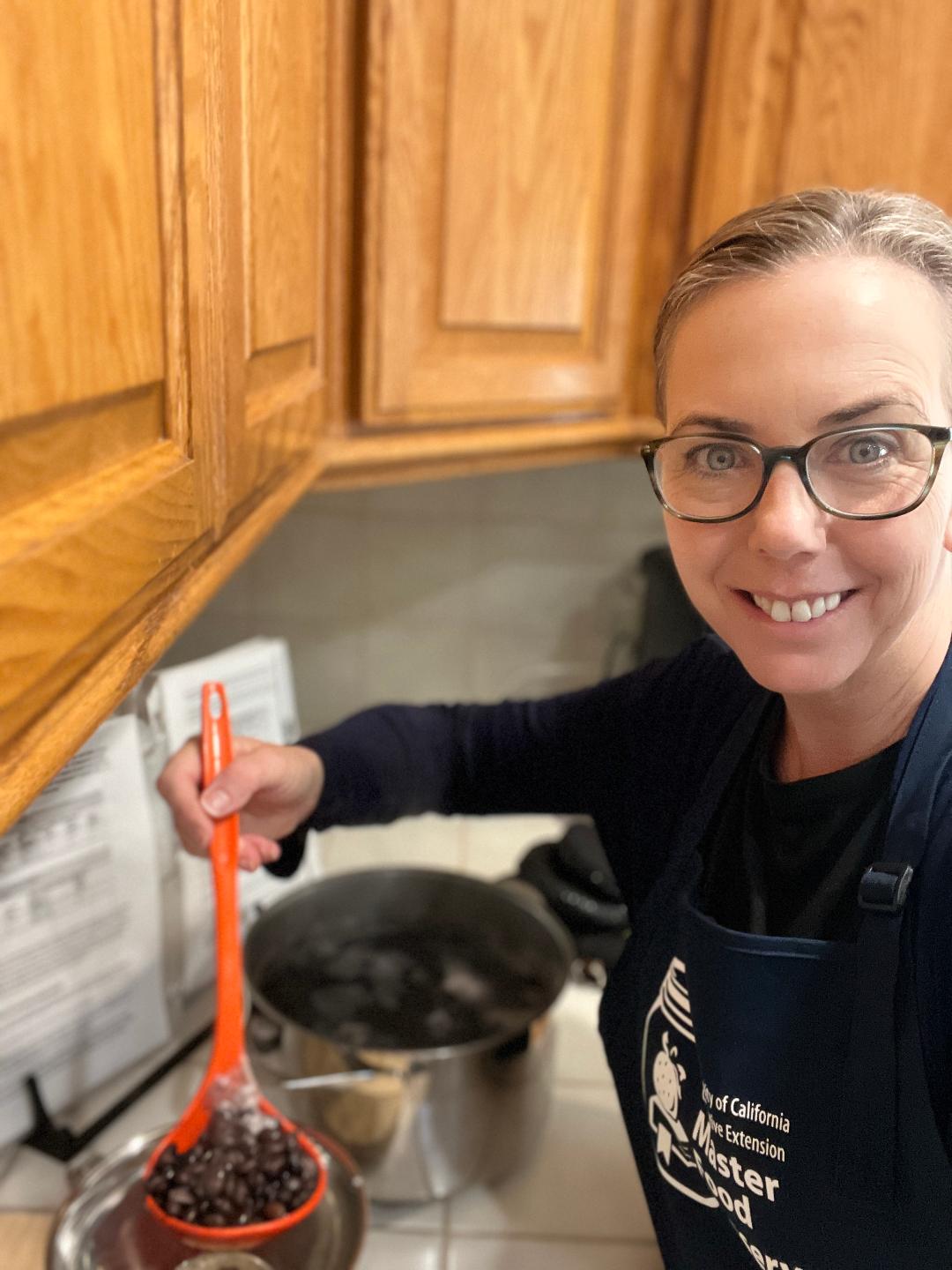 a woman wearing a Master Food Preserver apron in the kitchen ladling fruit into a pan