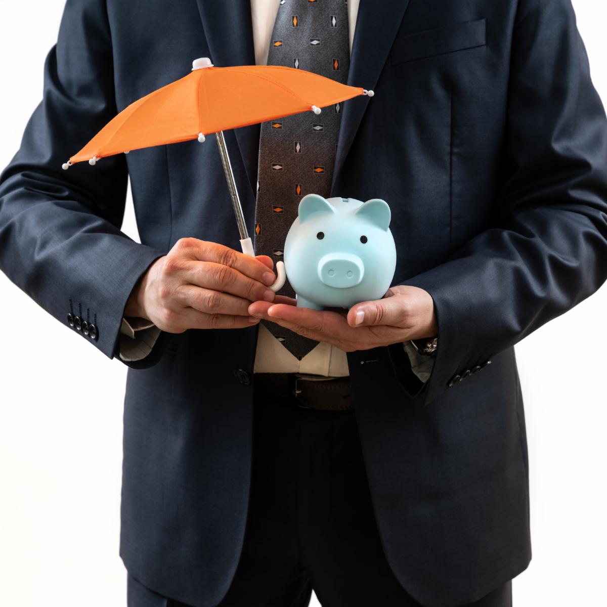 A man in a business suit holds a miniature umbrella protecting a piggy bank.