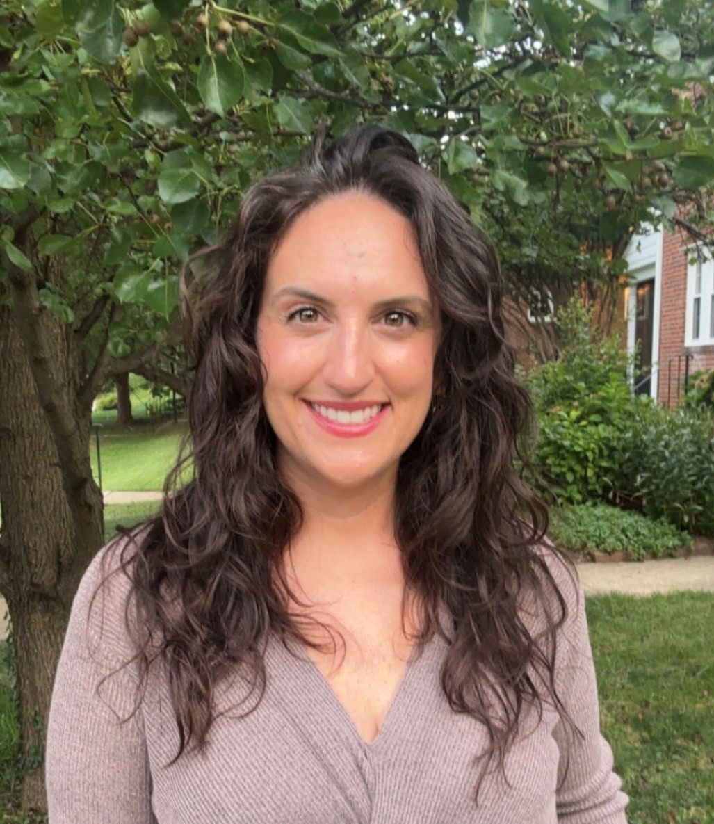 A person with long dark hair wearing a tan sweater in front of trees