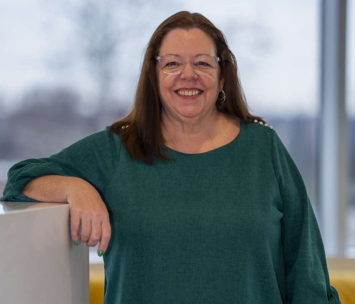Barbara Hoey smiling looking at the camera while wearing a green blouse, glasses and long brown hair