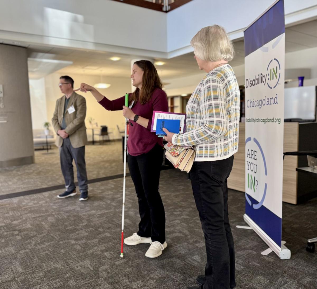 Photo of three people standing in an office with a Disability:IN Chicagoland banner behind them. The woman in the middle is speaking and holding a white cane.