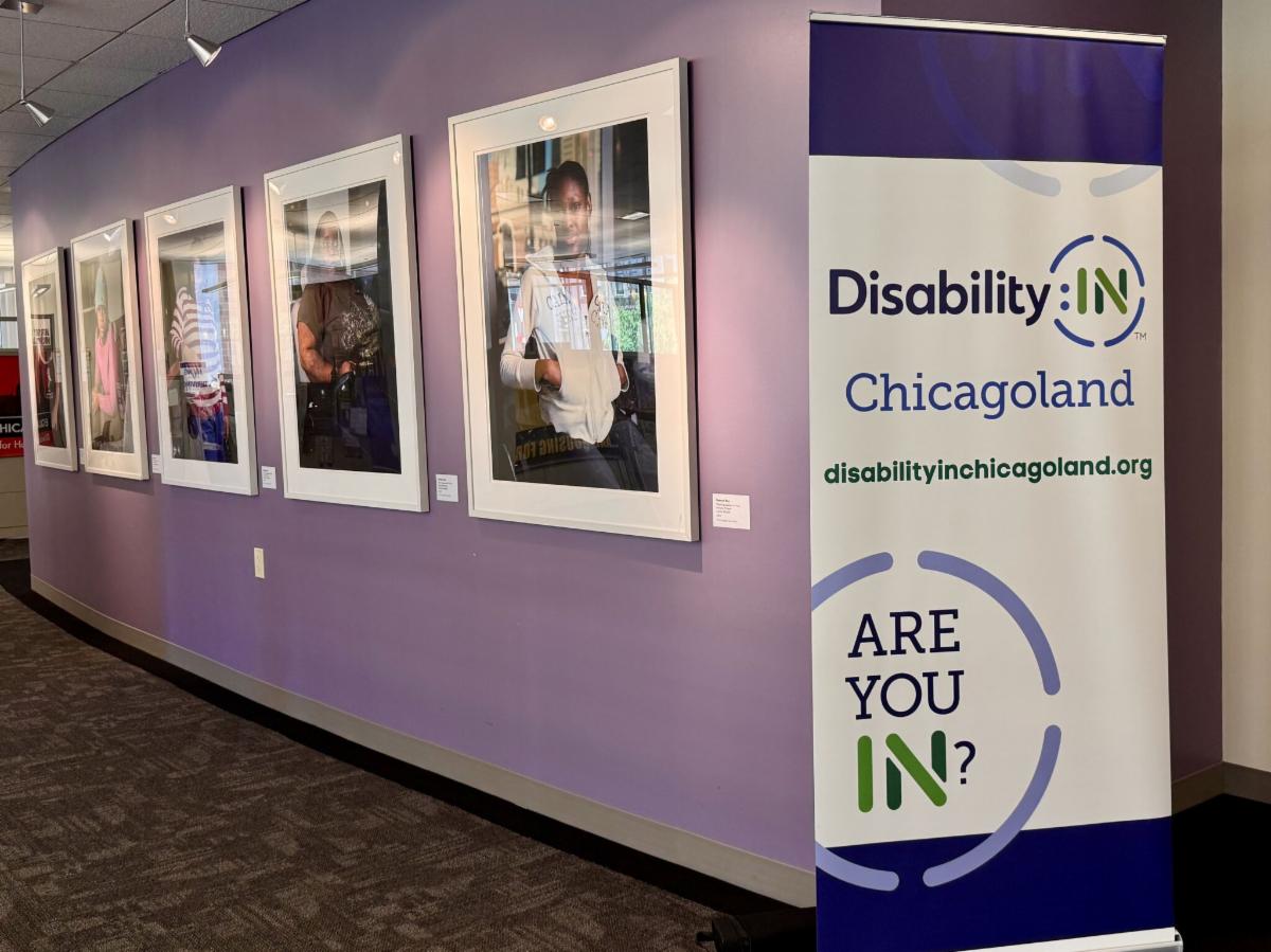 Photo of a Disability:IN Chicagoland banner with logo, website address and AreYouIN icon. The banner stands to the right of a series of pictures of individuals with disabilities hanging on a purple colored wall.