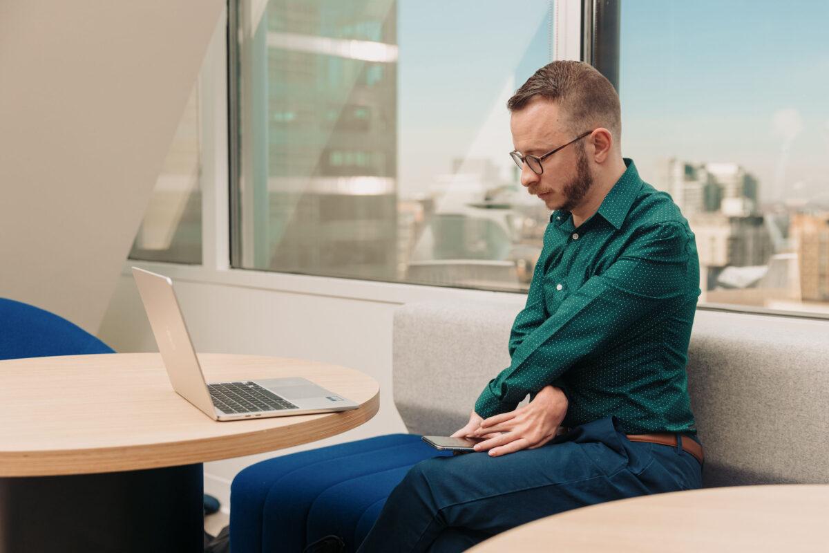 A man in an office works on a phone and a laptop. 