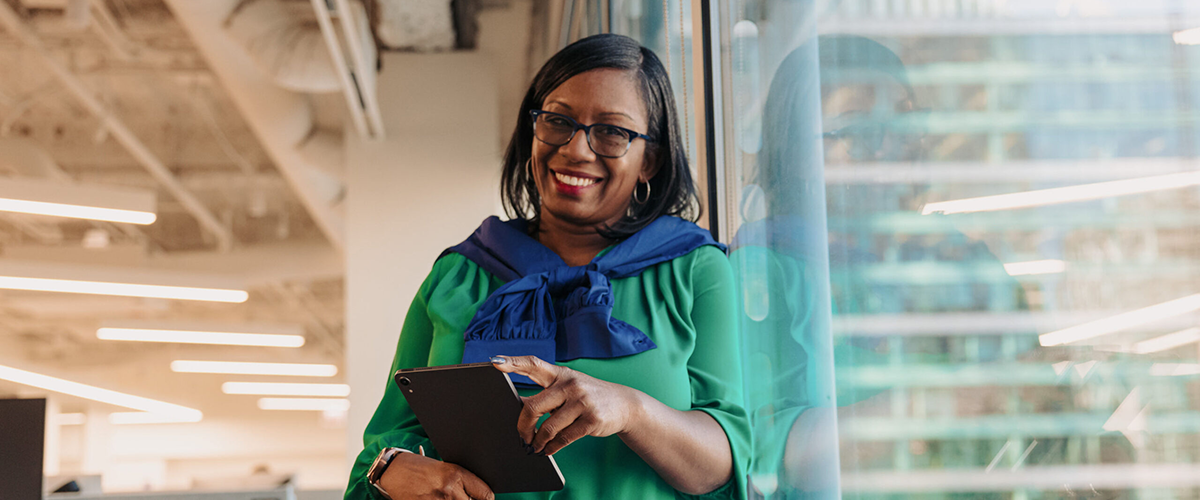 A woman with right side Hemiplegia smiles while leaning against a window in an office. She holds a tablet in her hands.