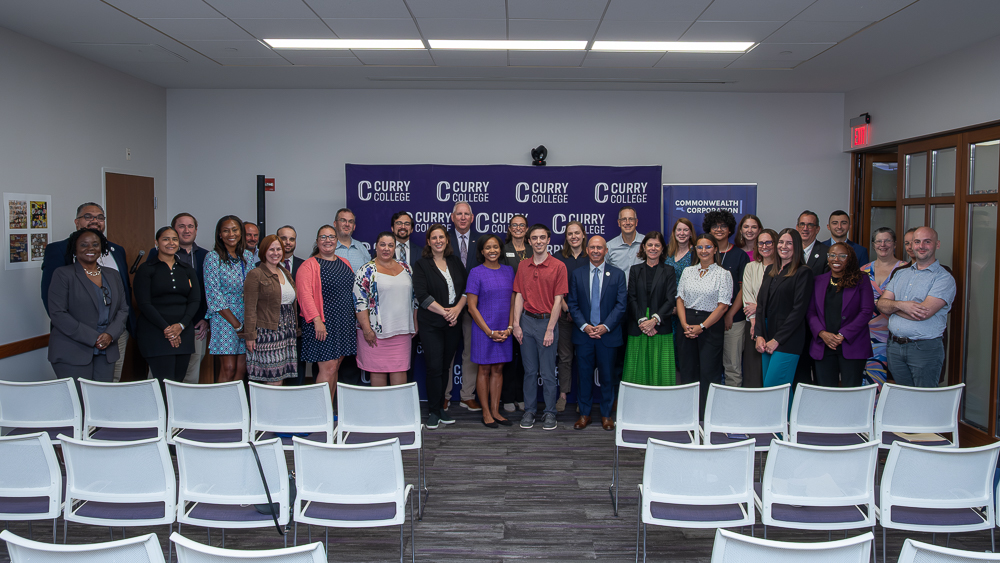 About 30 people pose for a photo in a classroom. Logos of Curry College in background.