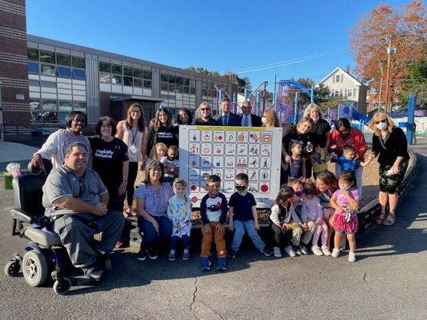 Group of children and adults gathered outside around a large communication board containing several icons. Playground and school building in background