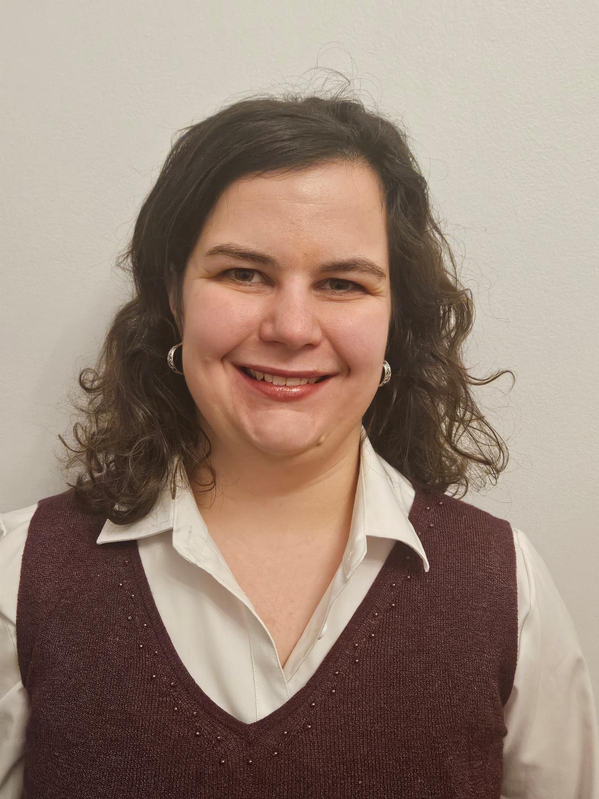 White woman with shoulder-length, curly brown hair wearing red sweater vest