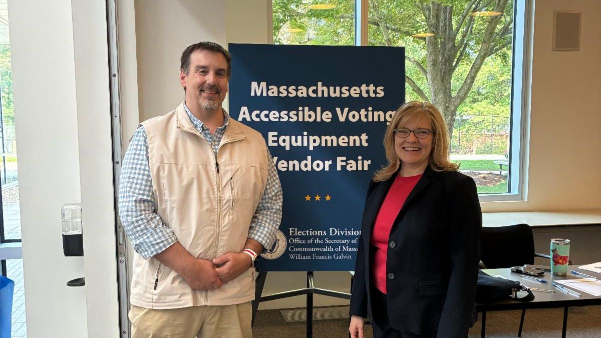 A man and woman stand in front of a sign that says Massachusetts Accessible Voting Equipment Vendor Fair